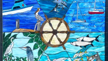 Stained-glass mosaic of ocean life and boats, with fish, birds, sea mammals, and sailboats arranged around a central ship’s wheel in vivid blues.