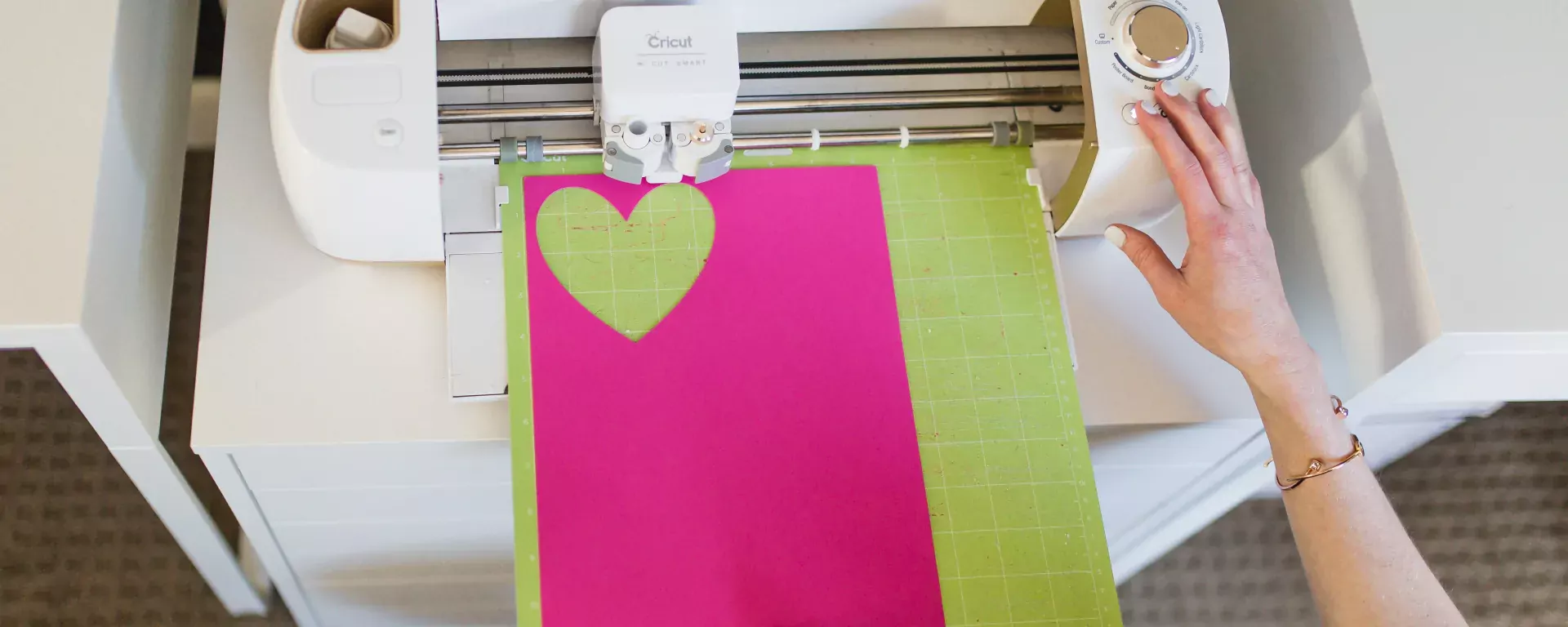 Photo of woman's hands inserting a cutting mat into a Cricut. Project on mat is a heart cut out of pink cardstock.