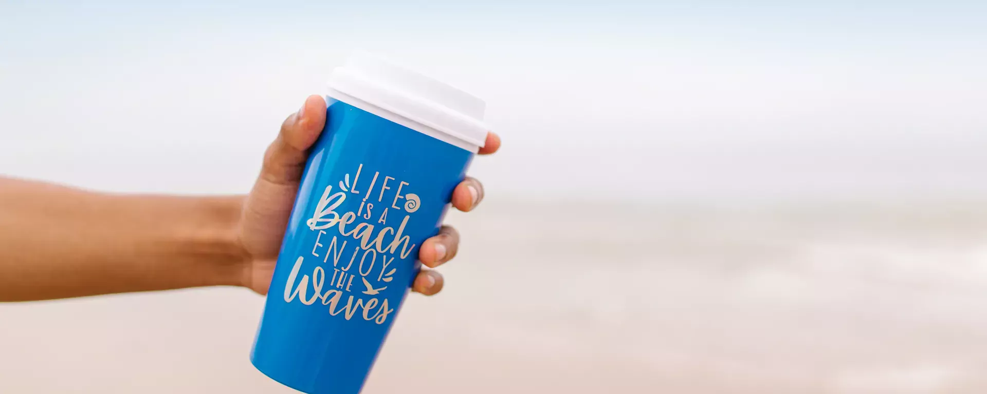 Close-Up Shot of Person Holding a Blue Tumbler at the beach. Text reads life is a beach, enjoy the waves.