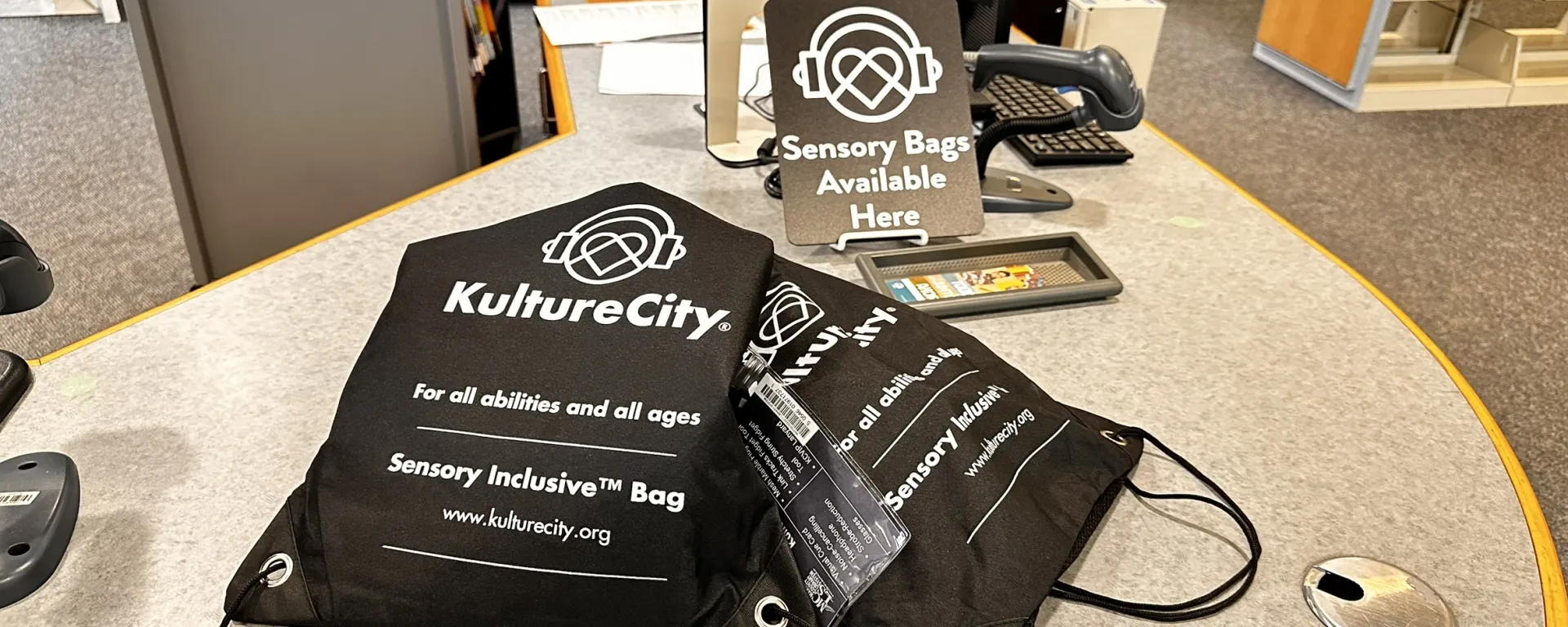 Two black canvas bags with "KultureCity" branding are on a library counter. A sign reads "Sensory Bags Available Here."