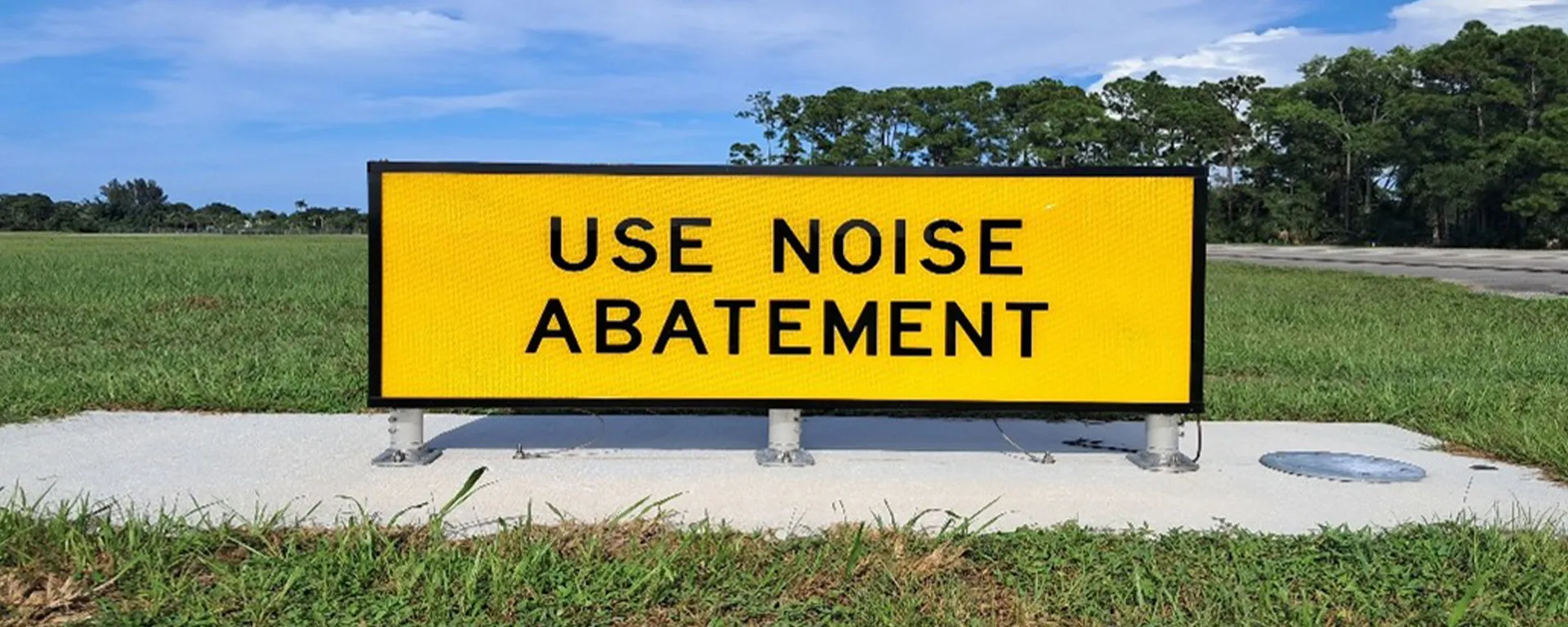 Yellow airfield sign reading “USE NOISE ABATEMENT” in black capital letters, mounted on a concrete pad beside a grass field.